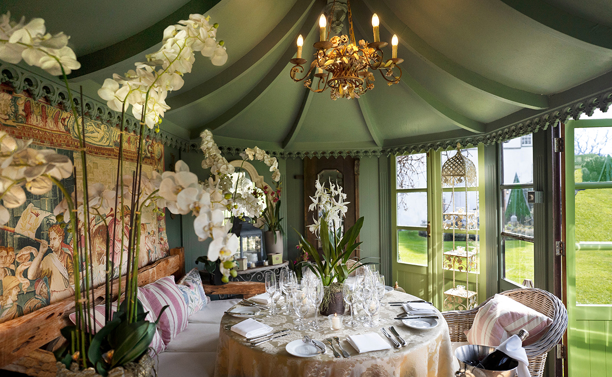 Beautifully furnished outdoor tea room with green painted ceiling and walls, colorful tapestry, chandelier, dining table, white orchid plants and many windows facing garden